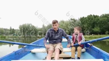 年轻快乐的父亲和他的儿子在绿色公园的木船上快速划船。 快乐的父亲和小孩一起在湖上度过时光